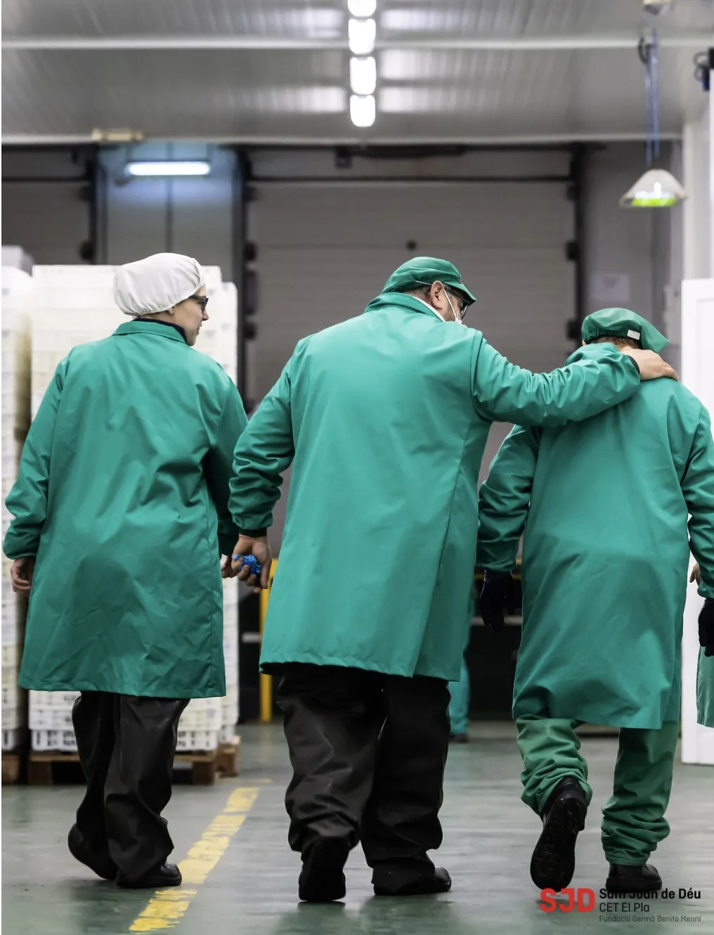Tres treballadors del CET El Pla amb bata i gorra verdes, d'esquena, caminant per la nau; un d'ells posa el braç sobre l'espatlla d'un company
